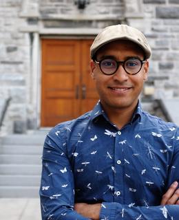 a brown man smiling directly at the camera, wearing a hat and wearing a blue shirt. 