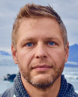 An image of Dr. Kit Dobson outdoors. He has short hair and a beard, and wears a blue sweater.