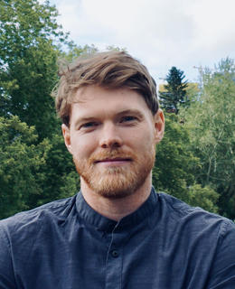 Headshot of Ben M. McKay wearing a dark blue shirt with trees in background.