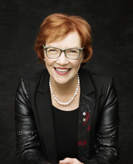 A photo of Dr. Aritha van Herk by Trudie Lee Photography. She has short red hair and glasses, and wears a black jacket and shirt, against a black background.