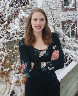 A woman outside is wearing a dark green floral shirt and black dress pants. Her hair is down and she has her arms crossed, looking at the camera. 
