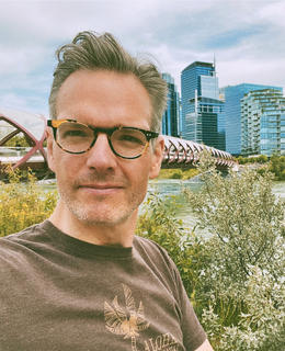 A photo of Dr. Michael Ullyot outdoors. He has short hair and glasses, and wears a brown shirt.