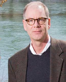 A headshot of Dr. Jim Ellis. He wears glasses, a brown blazer, and a navy-blue sweater with a white collared shirt.