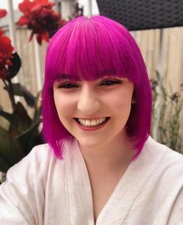 A wait-up photo of a person smiling at the camera. They have shoulder length, vibrant pink hair with bangs. They are wearing a white shirt. There are plants behind them. 