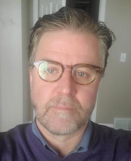 A photo of Dr. Jason Wiens. He has short hair and a beard, and wears glasses and a blue sweater.