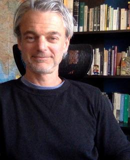 Smiling man with grey hair in black sweater, bookshelves in background