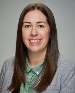 Photo of a white woman with brown hair, smiling, wearing a green shirt and white blazer.