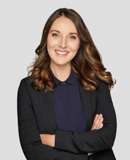 An image of Alexandra Handley. She wears a black blazer over a navy collared shirt. Arms crossed, she smiles at the camera.