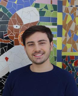 A picture of myself wearing a blue sweater. I am standing in front of a mosaic wall at Universidade Federal de Santa Catarina.