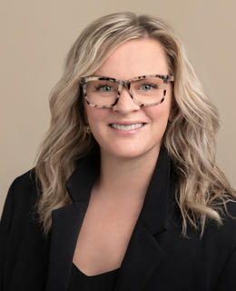 Dr. MacEachern has blonde hair and pale skin, and is wearing glasses and a black suit jacket
