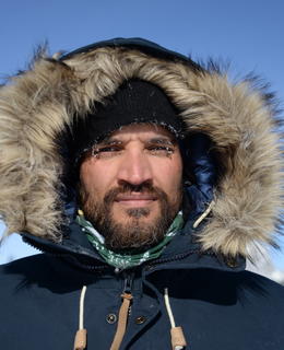 Headshot of Dr. Venkataraman in the field. He is wearing a fur-edged hood with snow-covered mountains in the background.