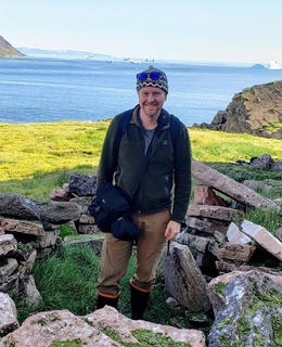 Photo of Mathew Walls standing at a fieldsite, the Archaeological Survey at Apparsuit, NW Greenland. He is surrounded by rocks with a field and oceans behind him. He is smiling at the camera.