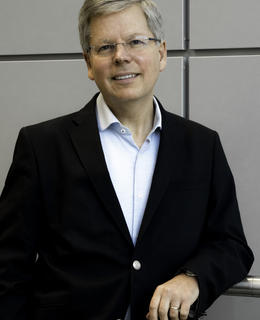 Image of male presenting person, wearing a dark suit, standing, leaning on ledge