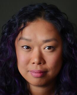Filipina woman with wavy black and purple hair. She is in front of a dark green background, smiling slightly.