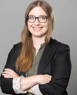 Julia smiles at the camera, she wears glasses, and has her arms crossed