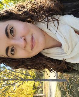 Christina has brown curly hair, brown eyes and wears a white scarf, green sweater and blue dress. She is facing forward and smiling at the camera.