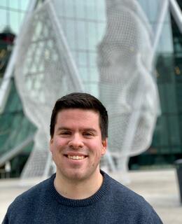 Alex is standing outside The Bow building in front of the Wonderland Sculpture. He has brown hair and blue eyes. He is wearing a dark blue shirt and is smiling. 