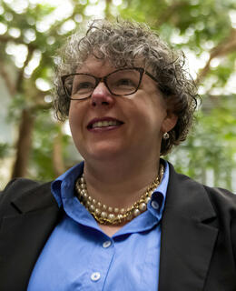 Sarah Elaine Eaton - A woman with short curly hair and glasses, wearing a blue shirt and a black jacket. The photo is taken outdoors and there is green foliage blurred in the background.