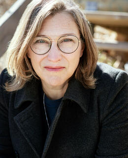 White woman, shoulder length brown hair, glasses, wearing a dark coat