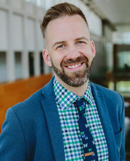 Image of Dr. Derritt Mason. They have short hair, a beard, and are wearing a blue jacket and blue and green checked shirt with a blue and green butterfly tie.