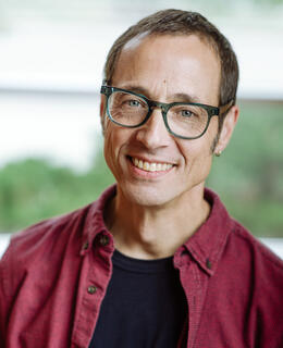A smiling man with short brown hair, a gold earring and tourquoise glasses.