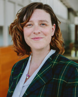 A white woman with short, wavy brown hair smiles at the camera wearing a plaid blazer and a white blouse.