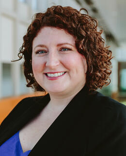 Image of a women with curly hair and wearing a black blazer and blue blouse