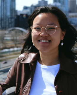 Photo of Ryanne, wearing glasses and a cool jacket, smiling in front of the Calgary cityscape.