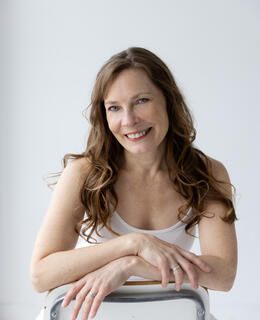 A smiling Claire leans on back of chair dressed in white.