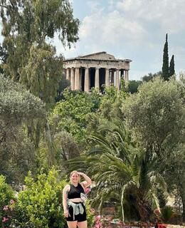 Sarah standing in front of the Temple of Hephaestus at the ancient Agora in Athens; Sarah is wearing a black top and shorts with a green sweater tied around her waist. Her left hand is raised to cover her eyes from the sun.
