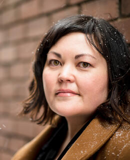 A photo of a mixed race Japanese Canadian woman looking at the camera. She has a short curly bob and is wearing a brown coat.