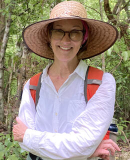 Photo of Kathryn Reese-Taylor in the forest. She is wearing a backpack and large straw hat and smiling at the camera.