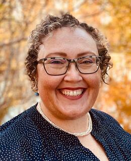 Dawn is shown with short blonde/grey, curly hair, and glasses. She is smiling in front of a background of fall leaves.
