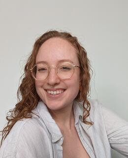 A young woman in her twenties smiles at the camera. She has long curly red hair and gold octagon glasses.