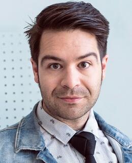 Anton deGroot's Headshot showing a clean shaven male presenting person in a jean jacket with a black tie on a light blue background. Very snazzy.