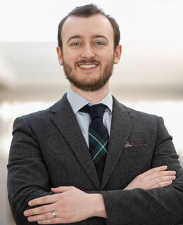Man with short brown hair and beard, blue eyes, wearing a tartan tie and grey blazer.