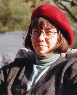 A woman with a short brown bob style haircut. She is wearing a red beret, thin black framed glasses, and a blue turtleneck shirt.