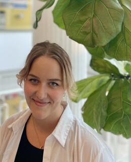 Young woman with blonde hair, white shirt, standing and smiling in front of a large plant.