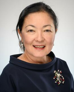 Professional headshot of Dr Yessenova wearing a dark blue top against a grey background. She is smiling.