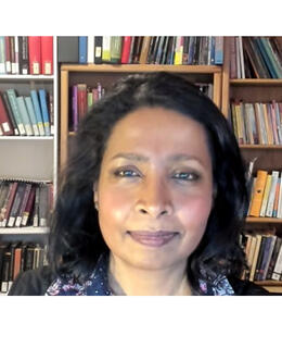 A photo of Clara A.B. Joseph. She has short black hair and wears a patterned navy-blue shirt, against a background of bookshelves.