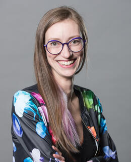 Headshot of Suzanna. She has long, brown hair, is wearing round, purple rimmed glasses, and a colourful blazer.