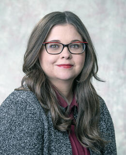 Cis white female with long brown hair wearing red and brown glasses, a burgundy blouse, and black and white marled cardigan