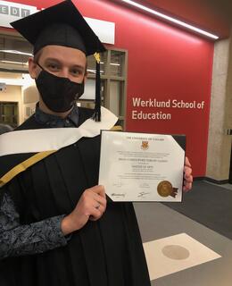Photo of Brian Gilbert wearing a mask and gown while holding up his MA degree outside of the Werklund School of Education