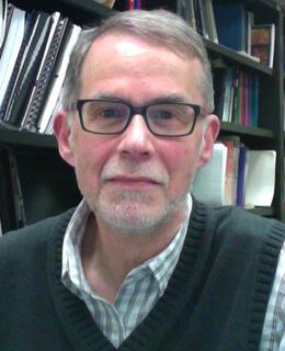 Portrait of a bespectacled Tom Langford with a short white beard, sitting in his office with shelves of books in the background