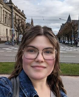 Image of a young woman with brown hair and glasses, buildings in the background.