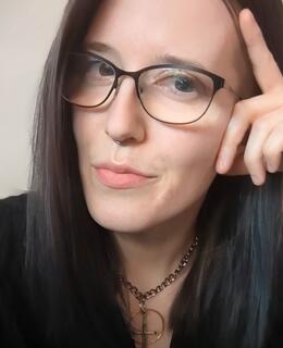 A headshot of J Overholser (a pale femme presenting individuals with black frame glasses and medium length dark hair) looking at the camera while leaning on one hand