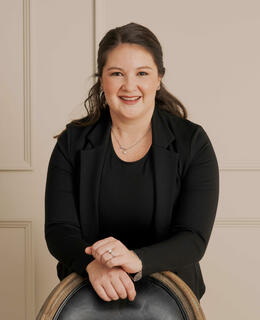 Heather Hinz's profile photo. She stands behind a chair with her hands crossed, leaning on the back of the chair. She is wearing a black blazer and a black dress.