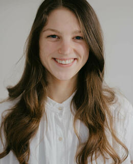 Image of Holly wearing a white shirt, smiling, with long brown wavy hair. 