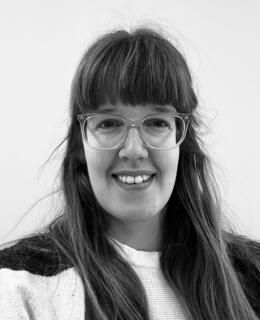 Black and white photo of Ellen looking directly into the camera. Ellen has long, dark blond hair, wears bangs and prescription glasses.