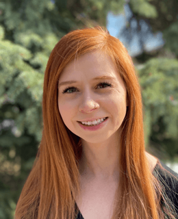 A close-up headshot of Kristina Waldmann smiling outdoors, with pine trees in the background